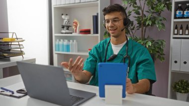 Tıbbi önlüklü genç bir İspanyol profesyonel ve modern iç mimari unsurlarla yapılan bir video görüşmesi sırasında klinikte kulaklık ve dizüstü bilgisayar kullanıyor..
