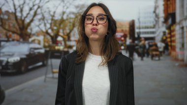 Londra 'da bir şehir caddesinde gülümseyen İspanyol kadın, gözlüklü ve beyaz gömlekli, şehir trafiği ve insanlarla çevrili..