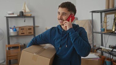 Genç adam yeni bir apartman dairesinde karton bir kutu tutarken akıllı telefondan konuşuyor. Etrafı eşyalarla çevrili, hareket sürecini sembolize ediyor..