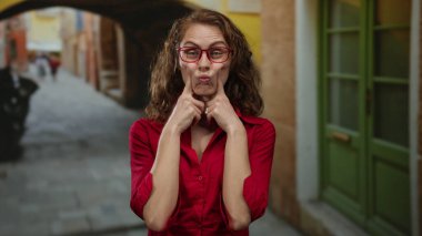 Gözlüklü genç bir kadın canlı bir şehir sokağında komik ifadeler takınıyor. Arka planda renkli binalar var. Kırmızı bir gömlek giyiyor..