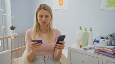 Bebek odasında beşik ve pelüş oyuncaklarla otururken akıllı telefon ve kredi kartı kullanan bir kadın, ev ortamında birden fazla işi ön plana çıkarıyor..