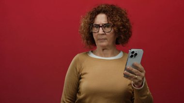 Gözlüklü ve kahverengi kazaklı bir kadın akıllı telefonu canlı kırmızı duvar arkaplanına karşı incelikle kontrol ediyor..