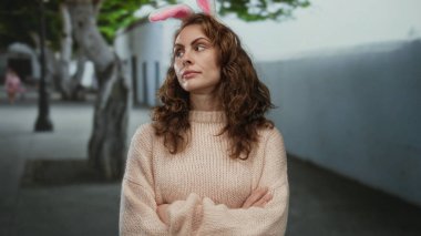 Tavşan kulaklı genç bir kadın şehir caddesinde kollarını kavuşturmuş, rahat bir kazak giyiyor, ağaçlı bulanık şehir geçmişine karşı ifade gösteriyor..