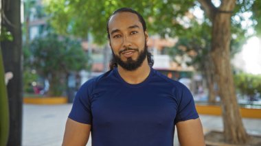 Şehrin arka planında güneşli bir günde şehir parkında mavi tişörtlü, sakallı genç bir Arap..