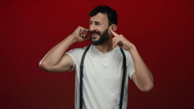 Kulakları kırmızı arka planda olan İspanyol adam beyaz gömlek pantolon askısı takıyor ve gürültü rahatsızlığını gösteriyor..