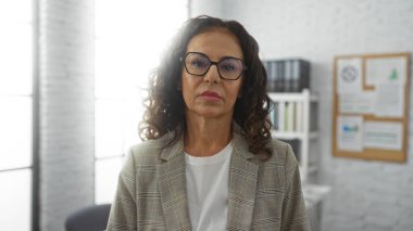 Gözlüklü kadın kendine güveniyor. Parlak bir ofiste. Ekose ceket giyiyor.