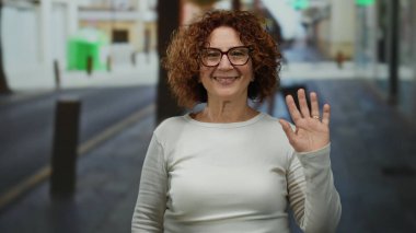 Güneşli bir günde esmer bukleleri ve gözlükleriyle şehir caddesinde gülümseyen kadın neşeli bir ruh hali sergiliyor..