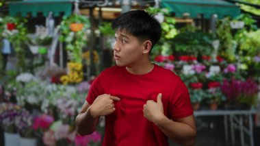 Kırmızı gömlekli genç adam şehirde canlı bir çiçekçi dükkanının önünde duruyor, renkli çiçeklerle çevrili, kalabalık bir sokakta ilginç bir anı yakalıyor..