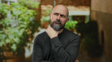 Sakallı, düşünceli, kel bir adam şehir merkezinde yeşil bir ortamda duruyor. Karar vermeyi düşünüyor.