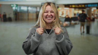 Havaalanı terminalinde gülümseyen, sarı saçlı, gri kazak giyen bir kadın, yolcuların hareket halinde olduğu canlı bir seyahat ortamı sergiliyor..