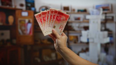 İsrail şekellerini tutan adam, finansal potansiyeli ve ekonomik büyümeyi vurgulayan görünür banknotların bulunduğu bir odada yatırımları ve iş fırsatlarını sembolize ediyor..