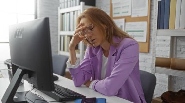 Modern ofis ortamında bilgisayar karşısında hayal kırıklığına uğrayan kadın, mor ceketli çekici genç kadın işyeri stresi, kitapların ve belgelerin olduğu kapalı bir ortam.