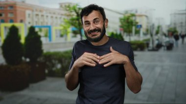 Arka planda bulanık binalar olan bir sokakta belirsizlik gösteren sakallı İspanyol bir adam..