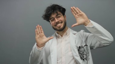 Gözlüklü, sakallı, beyaz önlüklü, kendine güveni ve dostluğu olan, gri bir arkaplana karşı gülümseyen ve el kol hareketi yapan genç İspanyol bir adam..