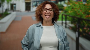 Esmer, orta yaşlı bir kadın arka planda yeşillik ve hareket halinde bir otobüsle şehir caddesinde gülümsüyor..