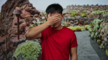 Kırmızı tişörtlü genç Çinli adam kayalık bir yolda dışarıda dikilirken elleriyle gözlerini kapatıyor..