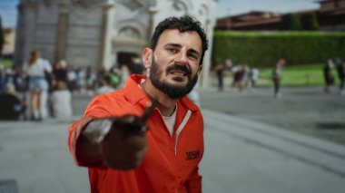 Turuncu tulumlu bir adam sidik kulesi ve arka plandaki insanlarla ön planda bıçak tutuyor ve şehir ortamında çarpıcı bir kontrast yaratıyor..