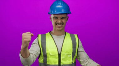 Yansıtıcı yelek ve kasketli genç adam, izole edilmiş pembe bir arkaplanda başarısını kendine güvenen bir jest ve gülümsemeyle ifade ederek kutluyor..