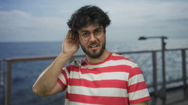 Sakallı ve gözlüklü genç adam yolcu gemisinde jest yapıyor. Arka planda okyanus manzarası, açık gökyüzünün altında kırmızı çizgili gömlek giyiyor.