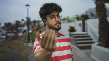 Genç, sakallı, İspanyol bir adam güneşli bir günde, arka planda palmiye ağaçları ve beyaz merdivenlerle gelmesi için el hareketi yapıyor. Rahat bir ortam izlenimi veriyor..