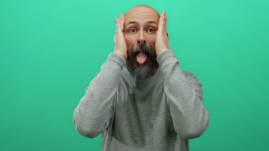 Sakallı kel adam izole edilmiş yeşil bir duvarın önünde neşeli tavırlarını ve günlük tarzını vurgulayarak neşeli ifadeler sergiliyor..