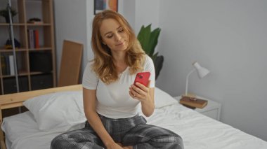 Modern yatak odasında beyaz dekorlu akıllı telefon kullanan, yatakta oturan, teknolojiyle uğraşan, rahat atmosfer, parlak ve davetkar ev ortamları olan bir kadın..