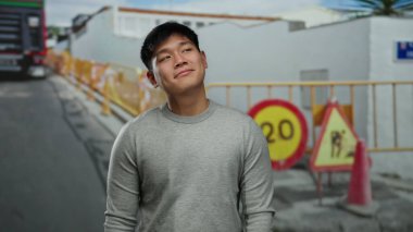 Açık hava inşaat sahasında duran gri kazaklı genç adam arka planda görünen tabelalar ve engellerle şehir altyapısını işaret ediyor..