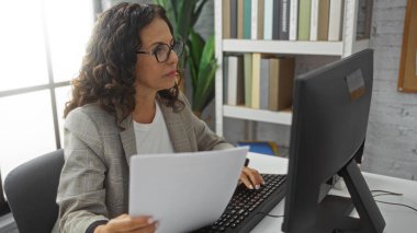 Modern bir ofiste çalışan bir kadın, elinde belgelerle bilgisayar kullanıyor, profesyonel bir çalışma ortamını resmediyor ve kapalı alandaki konsantrasyon ve verimliliğe odaklanıyor..