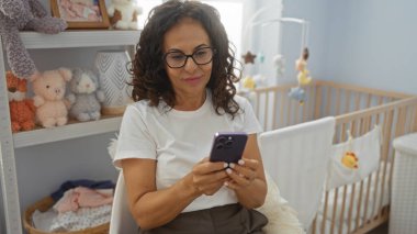Bebek odasında oyuncaklarla ve beşiğiyle oturmuş telefonla gülümseyen bir kadın, sıcak ve davetkar bir bağlantı ve konfor atmosferi yakalıyor..