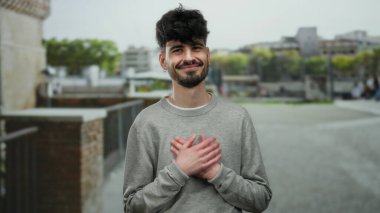 Sokak ortamında göğsünde elleri göğsünde sıcacık gülümseyen genç bir adam..