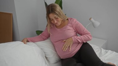 Yatak odasında sarı saçlı hamile bir kadın evde karnına dokunuyor. Huzur odasında bebeğini bekliyor..