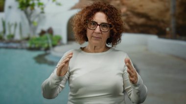 Elleriyle el kol hareketi yapan bir kadın açık havuzda yeşillik içinde.