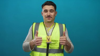 Gönüllü rozetli İspanyol asıllı genç adam, izole edilmiş mavi duvar arkaplanına ve toplum hizmeti ruhuna başparmak işareti yapıyor..