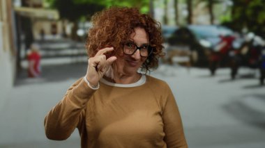 Woman gesturing small size on urban street with trees and blurred background, wearing glasses and brown sweater, demonstrating concept in city environment.