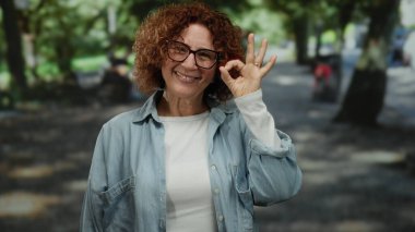 Yeşil ağaçlar ve açık hava ışıklarıyla çevrili, parkta gülümseyen bir kadın, huzurlu bir ortamda keyif ve güven anı yakalıyor..