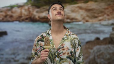 Genç İspanyol adam deniz kenarında dondurma yiyor, güneşli bir plaja bakıyor, açık deniz manzarasında kaygısız bir turist havası yaratıyor..