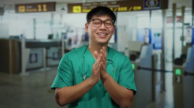 Yeşil önlüklü genç Çinli adam arka planda tabelaları olan kapalı bir havaalanı terminalinde alkışlıyor..