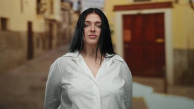 Beyaz tişörtlü, uzun siyah saçlı kadın şehir caddesinde gözleri kapalı duruyor. Gün ışığında huzurlu ve huzurlu bir ifade sergiliyor..