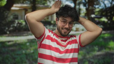 Gözlüklü, yeşil bir parkta kulak hareketi yapan, neşeli çizgili bir tişörtle güneşli ağaçların altında gülümseyen İspanyol bir genç..
