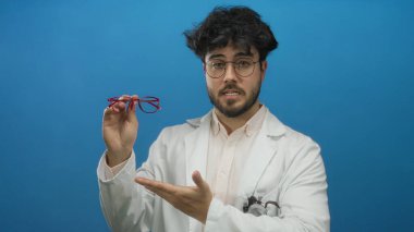 Sakallı, mavi arka planda kırmızı gözlük takan, steteskoplu beyaz laboratuvar önlüğü giyen, düşünceli bir yüz ifadesi sergileyen İspanyol genç adam..