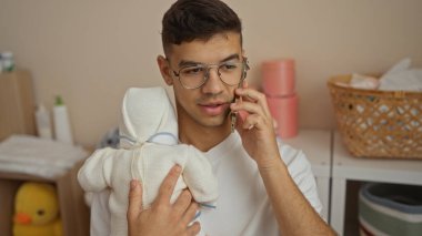 Bebek odasında beşik ve sepetlerle telefonda konuşurken kucağında bebek tutan genç bir adam aile hayatını rahat bir iç mekanda sergiliyor..