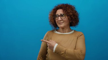 Orta yaşlı, kıvırcık saçlı, gözlüklü, parlak mavi arka planı olan, kahverengi bir süveter giyen orta yaşlı bir kadın..