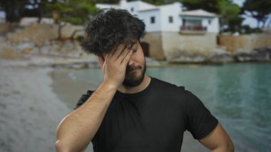 Deniz kenarında sakallı genç İspanyol bir adam gözleri kapalı bulutlu bir gökyüzünün altında plaj ve deniz zeminine karşı hüsran gösteriyor..