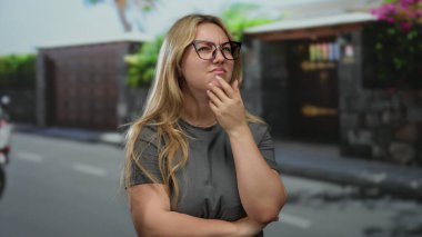 Açık havada, gözlüklü ve sarı saçlı bir kadın şehir caddesinde dikiliyor. Gri bir tişört giyerken tefekkür ediyor.