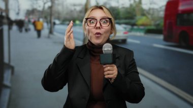 Elinde mikrofon tutan kadın bir şehir caddesinde şaşkın bir ifade ve arka planda bulanık trafik ve yayalar olan bir açık hava haberi sırasında.
