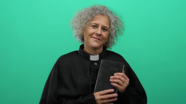 Gri saçlı bir rahip gibi giyinmiş bir kadın, izole edilmiş yeşil bir zemine karşı İncil 'i tutuyor, kameraya sıcak bir şekilde gülümsüyor, inanç ve maneviyatı somutlaştırıyor..