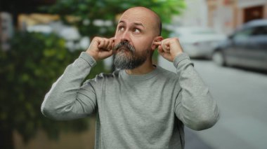 Gri kazaklı kel bir adam şehir sokağında dikiliyor, kulaklarını parmaklarıyla kapatıyor, arka planda bulanık yeşillik ve arabalar var, hayal kırıklığı ya da gürültü ifade ediyor..