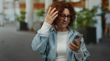 Açık havada akıllı telefon tutan kadın, şehir arka planı ve gündüz sahnelerinin yer aldığı hareketli sokak ortamında alnı ve yorgunluğu yüzünden endişeli görünüyor..