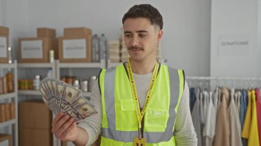 Yansıtıcı yelek giyen genç adam bize dolar veriyor. Arka planda kutular ve kıyafetlerle bir hayır kurumunda gönüllü olarak çalışıyor..