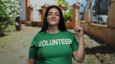 Yeşil gönüllü tişörtü giymiş iri yarı bir kadın güneşli park ortamında yukarıya doğru bakıyor. Açık havada gönüllülük ve toplum ruhunu sergiliyor..